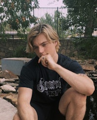a young man sits on a rock next to a pond